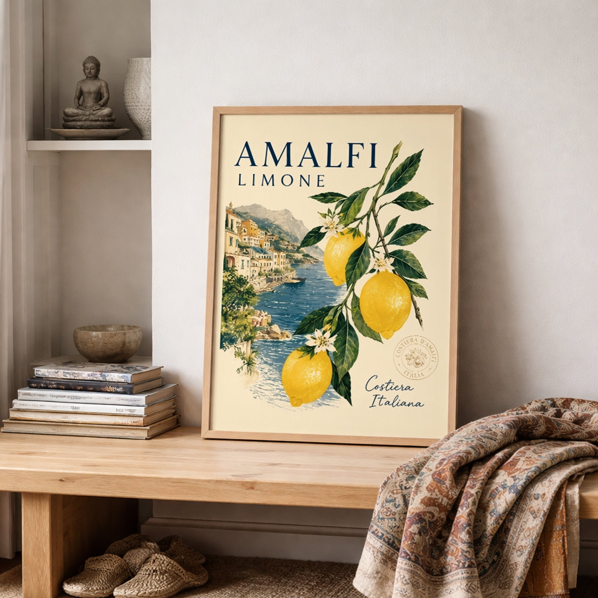 Framed Amalfi Coast lemon wall art styled on a shelf with books, soft fabric and neutral room decor. The Italian poster shows yellow lemons, green leaves, blossom flowers and a coastal sea view, creating a bright Mediterranean travel print for relaxed summer interiors.