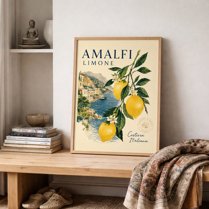 Framed Amalfi Coast lemon wall art styled on a shelf with books, soft fabric and neutral room decor. The Italian poster shows yellow lemons, green leaves, blossom flowers and a coastal sea view, creating a bright Mediterranean travel print for relaxed summer interiors.