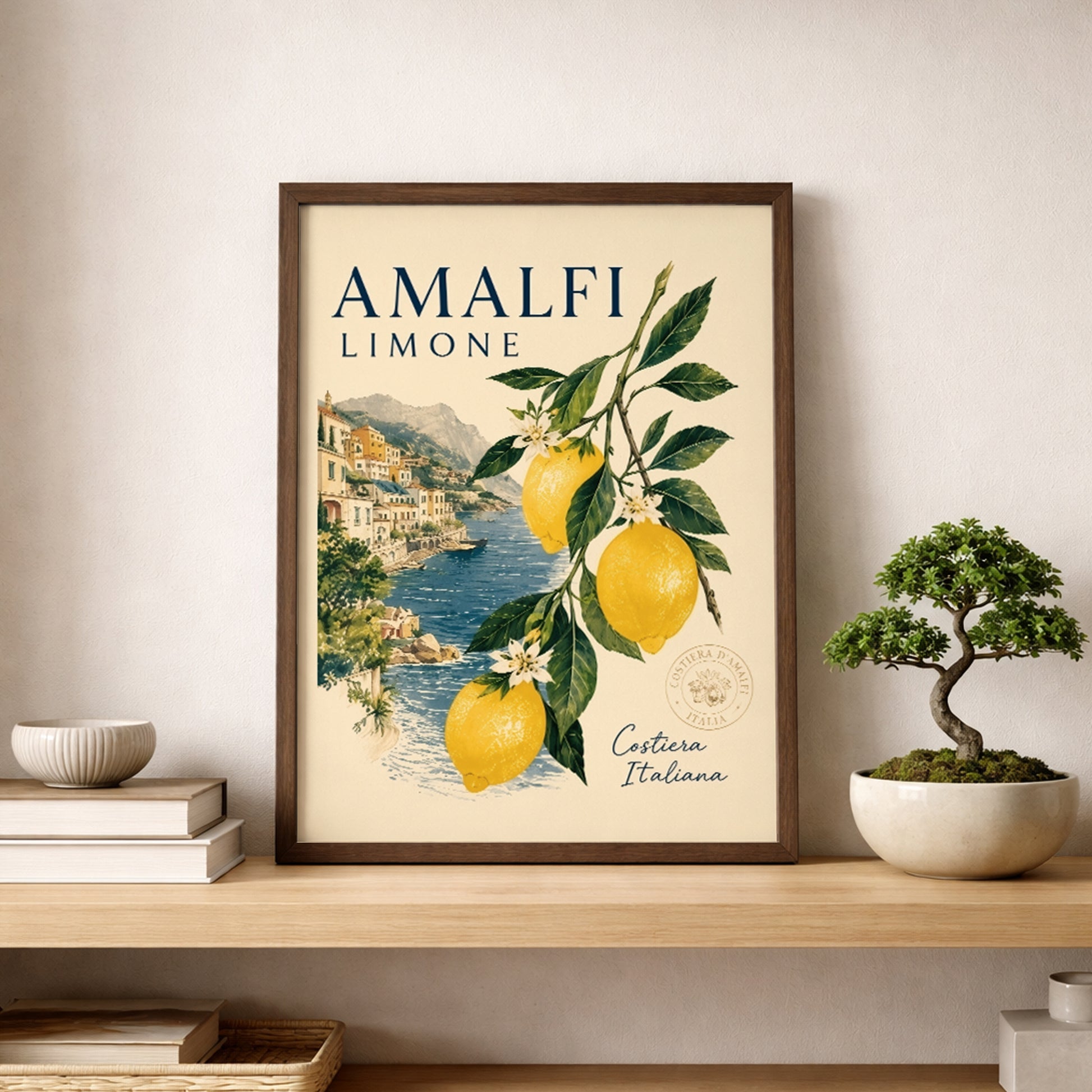 Amalfi Coast lemon poster in a wooden frame placed on a shelf beside a small bonsai plant and neutral home decor. The print shows yellow lemons, green leaves and an Italian seaside village, making it a fresh Mediterranean wall art piece for kitchen, dining or living spaces.