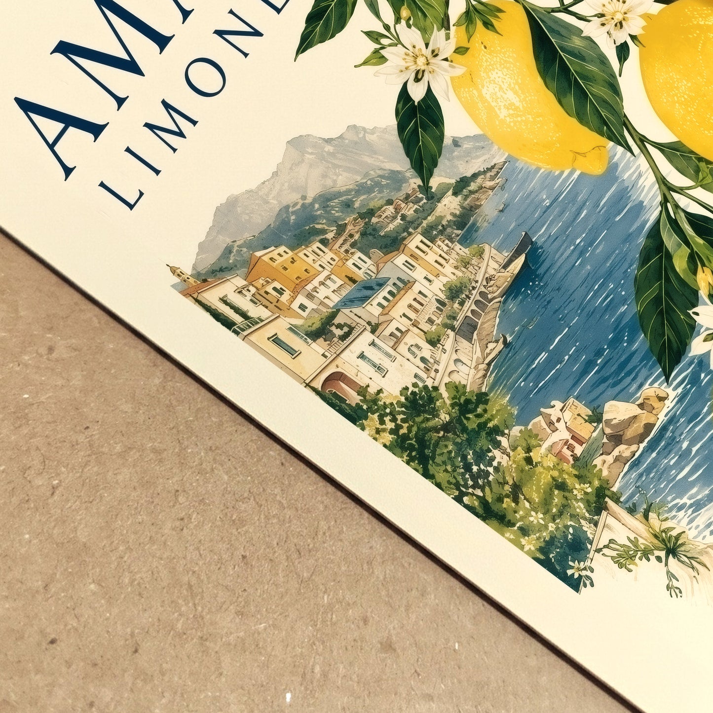 Close up of an Amalfi Coast lemon poster lying on a textured surface, showing the printed paper detail, yellow citrus fruit, green leaves, blue sea and Italian village artwork. A vintage style Mediterranean travel print with fresh summer colour and coastal character.