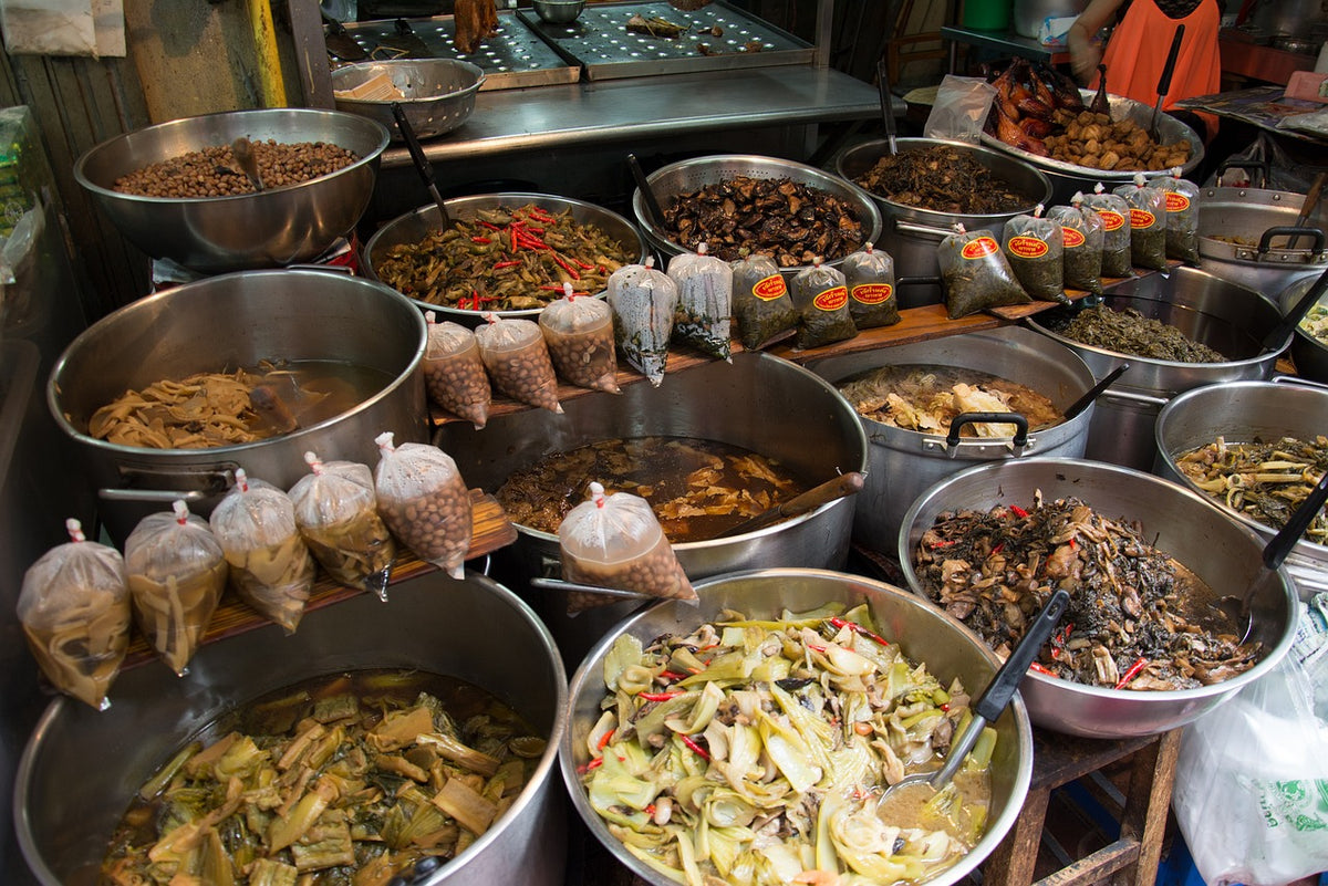 Prepared foods, pickled vegetables and packaged ingredients displayed in large metal bowls at a traditional Thai market stall.