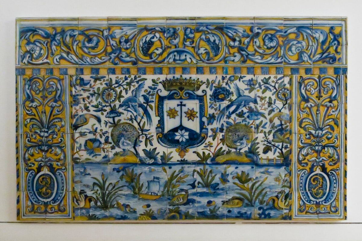 A square ceramic tile wall panel featuring a detailed hand-painted Portuguese style design with a central coat of arms, birds, foliage, and a blue water scene, decorated with an ornate blue and yellow border.

Manuelvbotelho, CC BY-SA 4.0 <https://creativecommons.org/licenses/by-sa/4.0>, via Wikimedia Commons

https://commons.wikimedia.org/wiki/File:Frontal_de_altar_azulejos_sec_XVII_IMG_1221.JPG