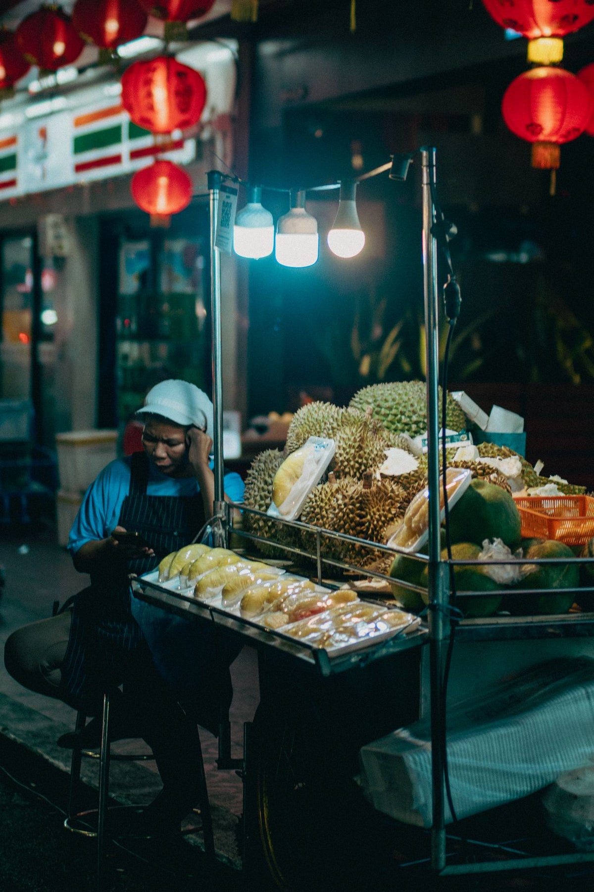 Thai street vendor selling fresh durian at a night market, with whole durians and prepared fruit displayed under market lights.