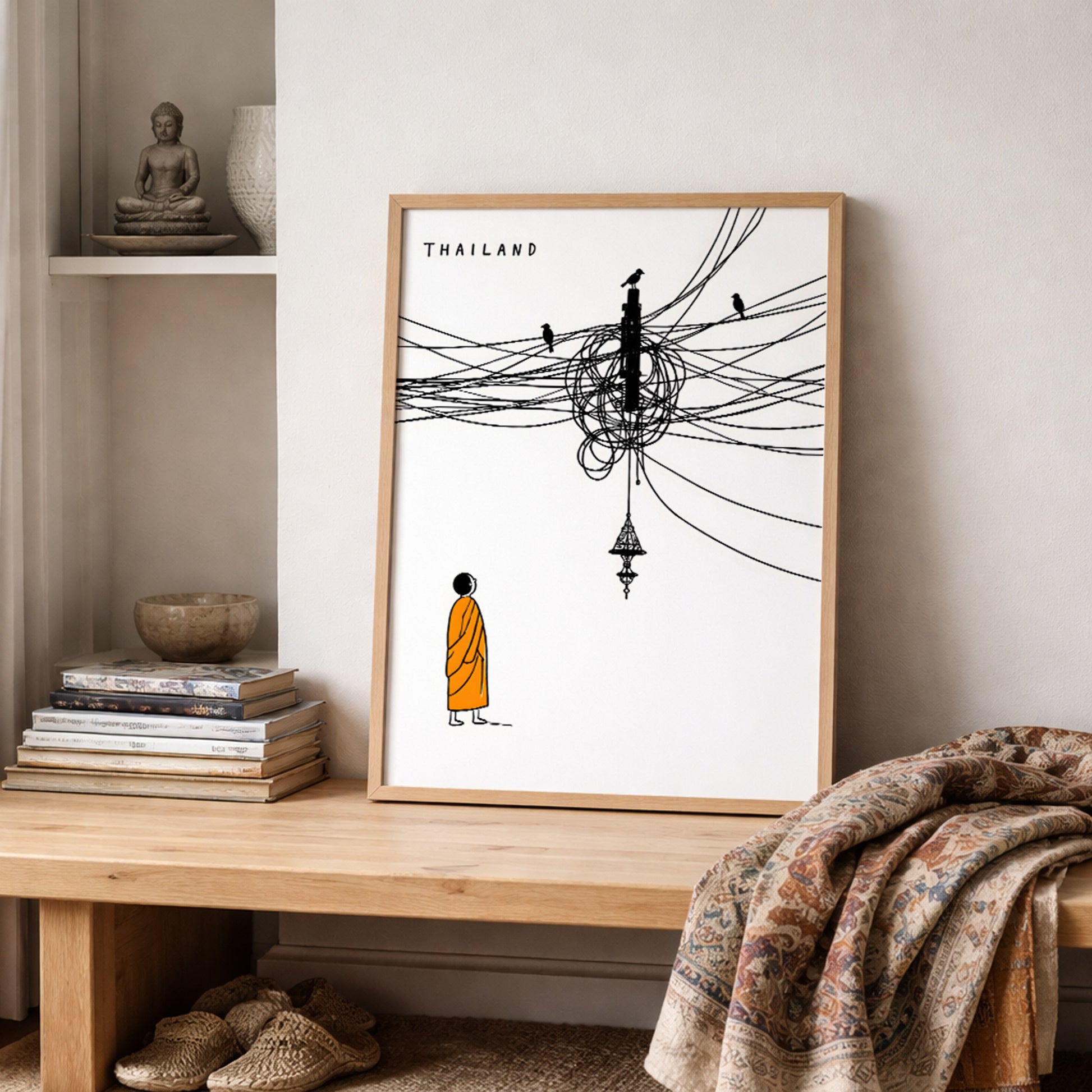 Thailand cable mess poster styled on a wooden shelf beside folded fabric and neutral home decor. The print shows a Buddhist monk in an orange robe below tangled black street cables, birds and a hanging Thai lamp, giving the room a clean travel art and cultural wall decor feel.