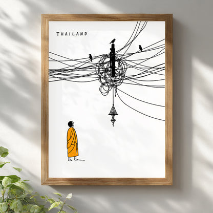 Wooden framed Thailand cable mess poster leaning against a bright white wall with soft natural light and greenery nearby. The artwork shows a small Buddhist monk in orange below tangled black overhead wires, birds and a Thai hanging lamp for clean cultural travel wall art.