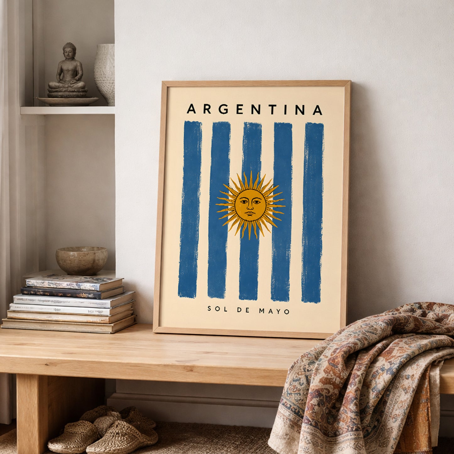 Argentina flag poster leaning on a wooden shelf in a softly styled neutral interior. The print features blue and cream vertical stripes, a golden Sun of May and Argentina lettering on a warm off-white background. Folded textiles, books and understated decorative objects create a relaxed setting for this Argentine cultural wall art.