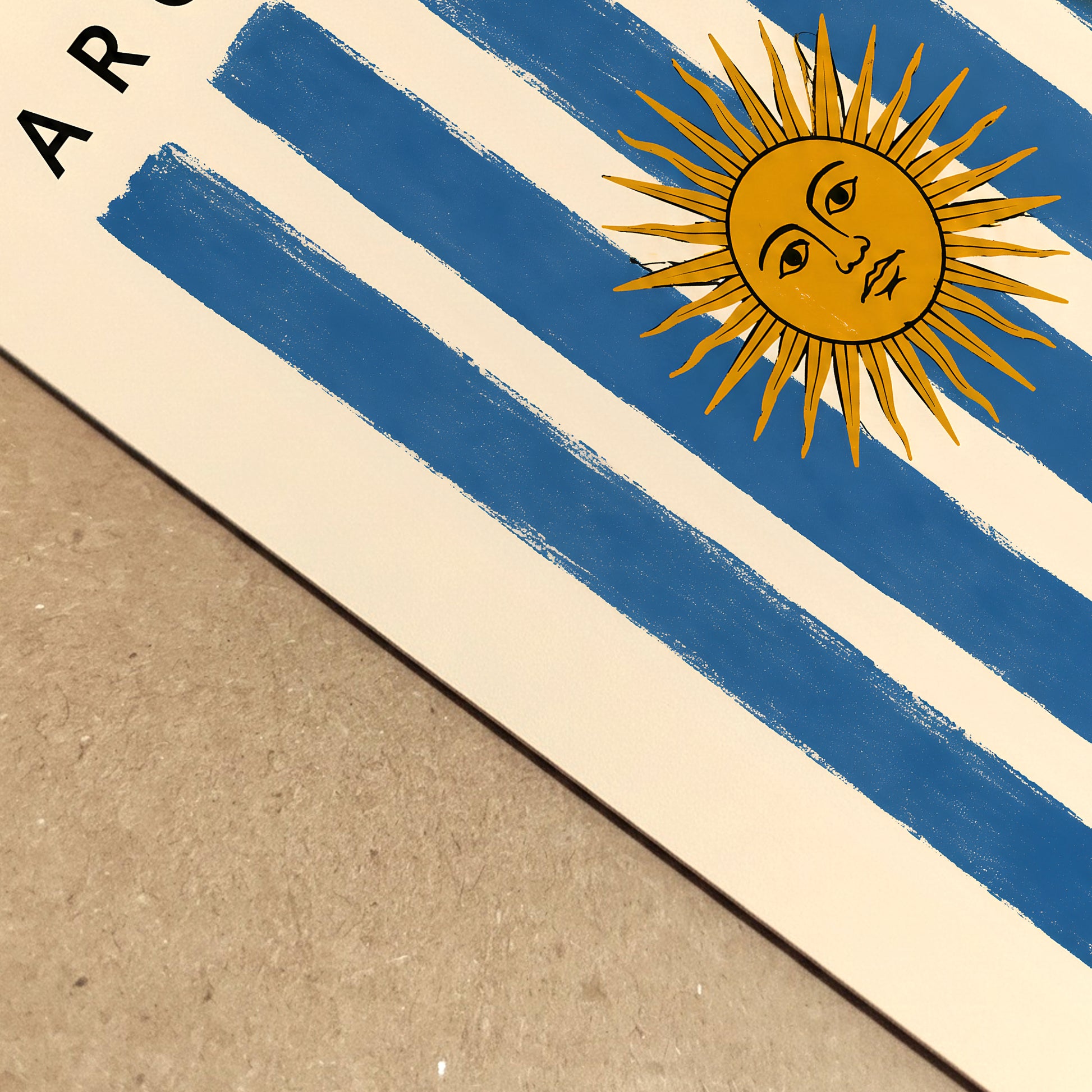 Close-up view of an Argentina flag poster resting on a natural brown surface, showing the printed paper and layered edge in detail. Blue and cream stripes lead towards the golden Sun of May beneath partially visible Argentina lettering. The angled composition highlights the texture, colour and vintage character of the Argentine print.