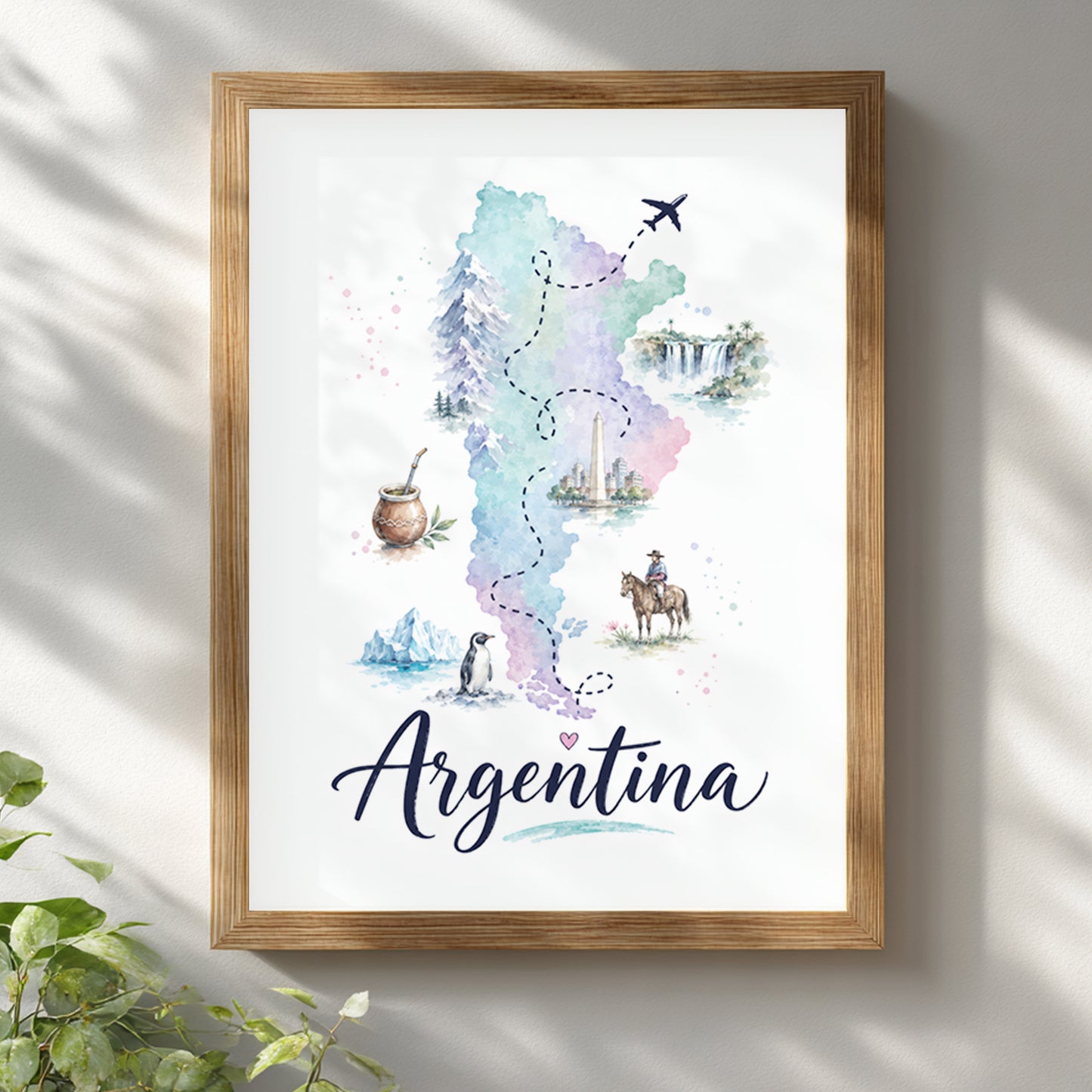 Argentina watercolour map poster displayed in a light wooden frame on a bright white wall with soft natural shadows. The artwork combines a pastel map of Argentina with small wildlife, travel and cultural illustrations, finished with large cursive Argentina lettering. Green leaves at the edge add a fresh, relaxed home decor setting.