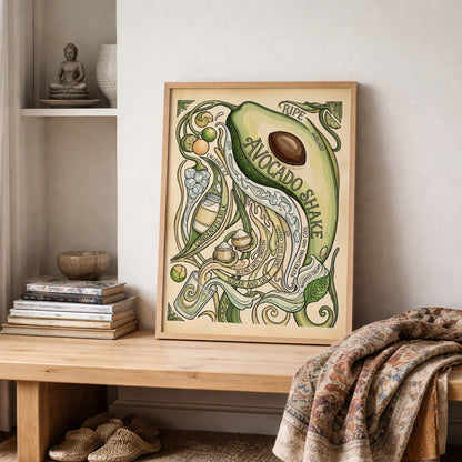 Framed avocado shake poster placed on shelving in a neutral home setting with stacked books, pottery and folded textiles. The artwork combines a large avocado, flowing vines, lime and recipe elements in a soft green palette. Styled as decorative kitchen poster art, it suits food wall art displays, tropical interiors and relaxed gallery wall arrangements.