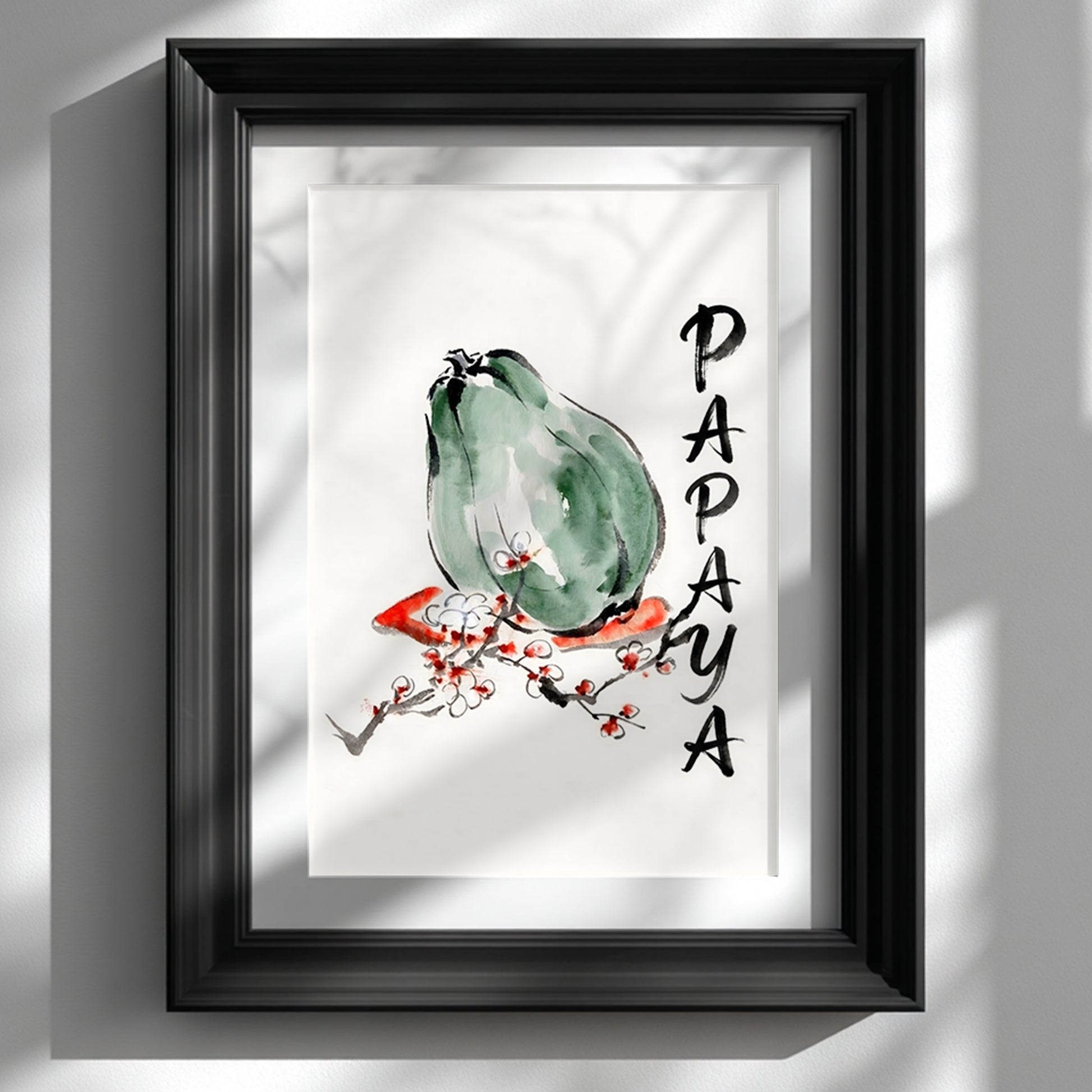 Framed papaya wall art shown in a black frame on a white wall with soft window shadows. The print features a green papaya in loose ink and watercolour style, a red and white blossom branch, and vertical hand painted PAPAYA lettering. Minimal tropical fruit wall art for kitchen decor, dining room styling, modern Asian inspired interiors, and clean botanical gallery walls.