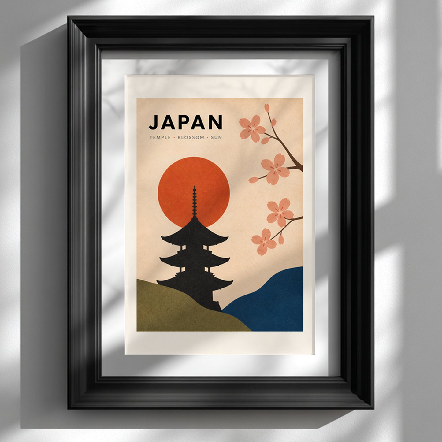 Vintage Japan travel poster shown in a deep black frame against a clean white wall. The artwork has a black Japanese temple silhouette, red rising sun, pink cherry blossom branches, olive green and blue hills, and a warm cream background, creating bold retro Asian wall art for a living room, bedroom or office.