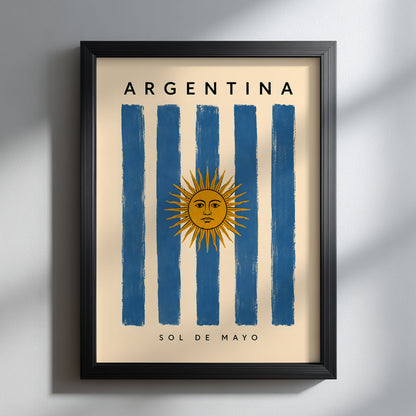 Argentina flag poster presented in a slim black frame against a softly lit grey wall. The vertical artwork combines blue and cream stripes with the golden Sun of May, Argentina lettering and Sol de Mayo text. Strong natural shadows give this Argentine travel print a clean, contemporary appearance suited to neutral interiors.