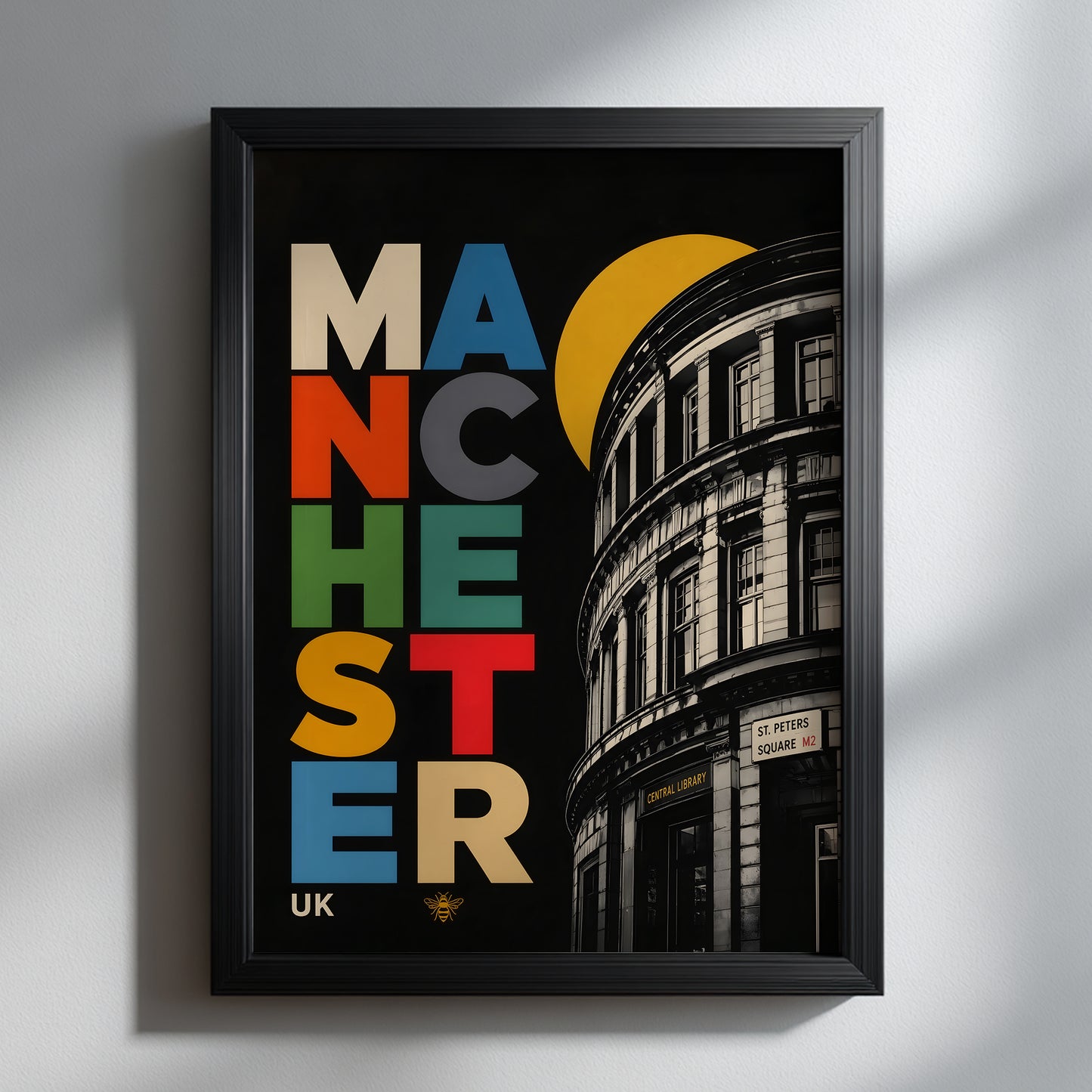 Bold Manchester Central Library wall art in a slim black frame on a light grey wall, showing colourful stacked typography, a yellow circle and monochrome curved architecture.