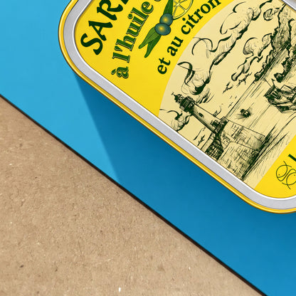 Angled close-up of a French sardine tin pop art print showing the paper surface, turquoise blue background and detailed yellow vintage food label with French lettering, coastal imagery, olives and lemon.