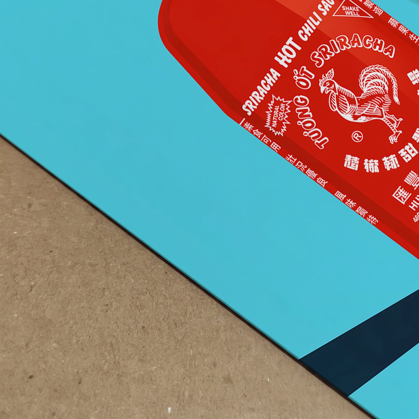 Close-up detail of the sriracha poster print, showing the red sauce bottle artwork, white label text, bright blue background, clean print edge and strong diagonal shadow. A bold pop art food print with colour depth and texture, made for kitchen decor, cafe wall art, restaurant styling or foodie interiors.
