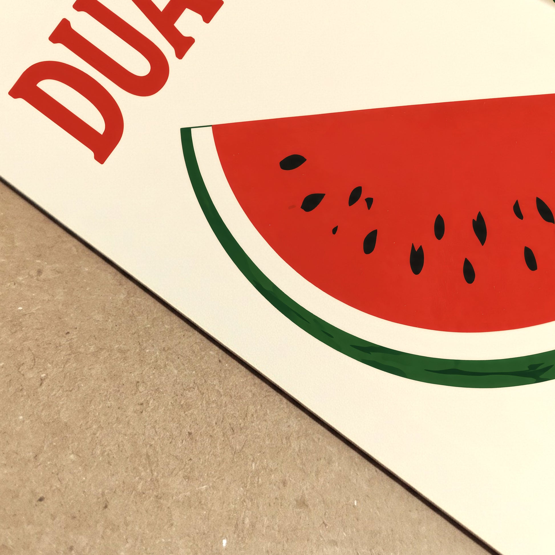 Close-up of a Vietnam watermelon poster print showing the edge of the paper, cream background, red DƯA HẤU lettering and part of the bold watermelon artwork. The angled view highlights the printed texture and retro fruit poster style, useful for kitchen decor and food wall art listings.