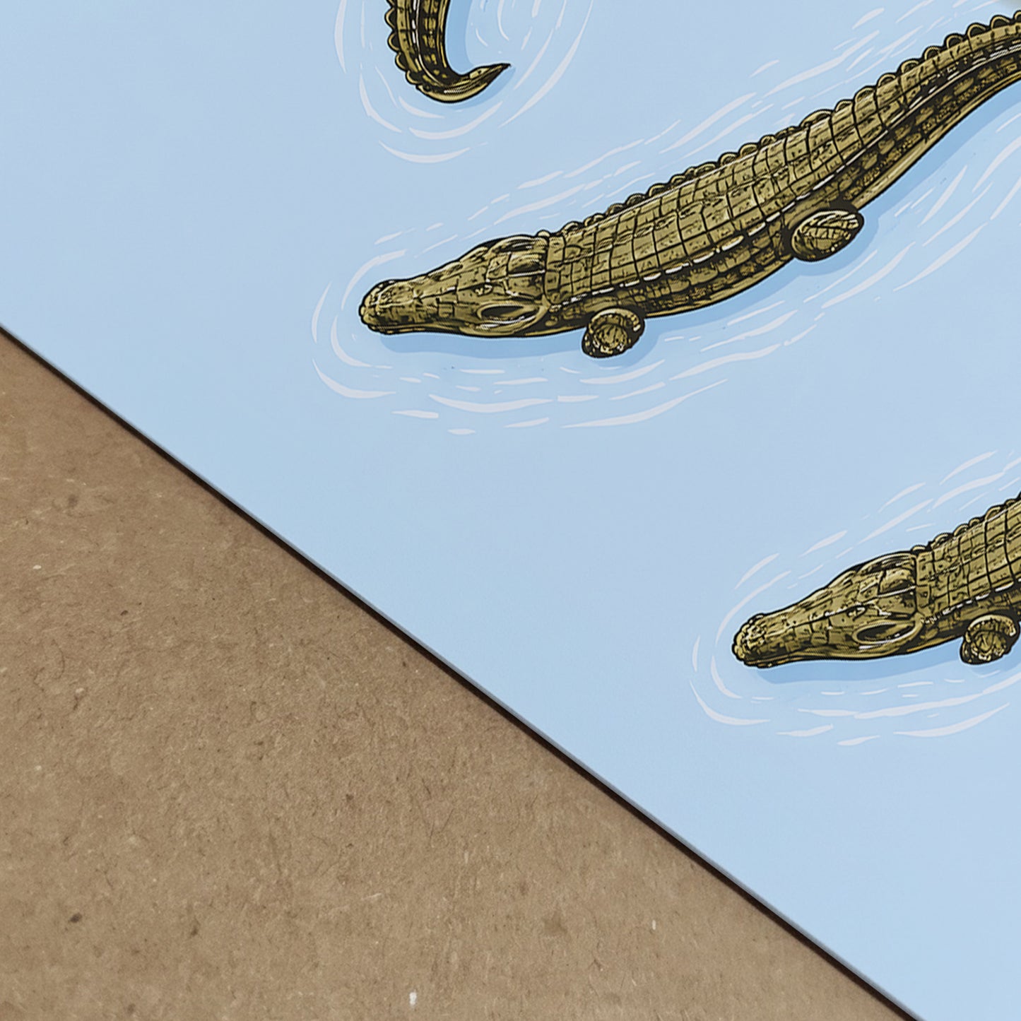 Close-up photograph of the crocodile art print showing paper texture and crisp illustration detail. The blue background meets a neutral surface, emphasising the fine woodcut-style scales and white water ripple lines around the reptiles. A detailed natural history reptile poster designed for wildlife wall art and zoology inspired decor.