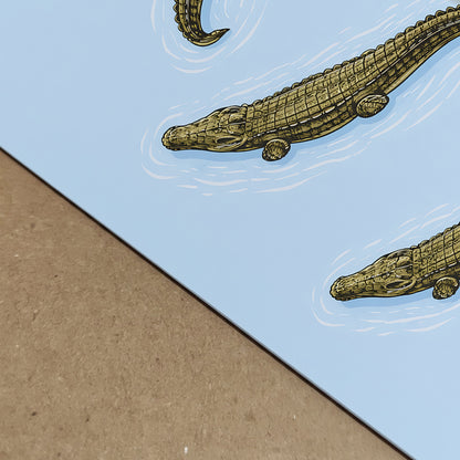 Close-up photograph of the crocodile art print showing paper texture and crisp illustration detail. The blue background meets a neutral surface, emphasising the fine woodcut-style scales and white water ripple lines around the reptiles. A detailed natural history reptile poster designed for wildlife wall art and zoology inspired decor.