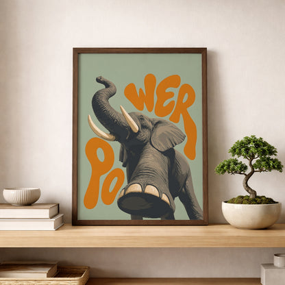 Elephant wall art displayed in a wooden frame on a shelf beside a small bonsai plant, showing a grey elephant with raised trunk, tusks and orange retro text on a soft green background. A bold animal wall print for calm living room decor, natural interiors, office styling or modern safari poster displays.
