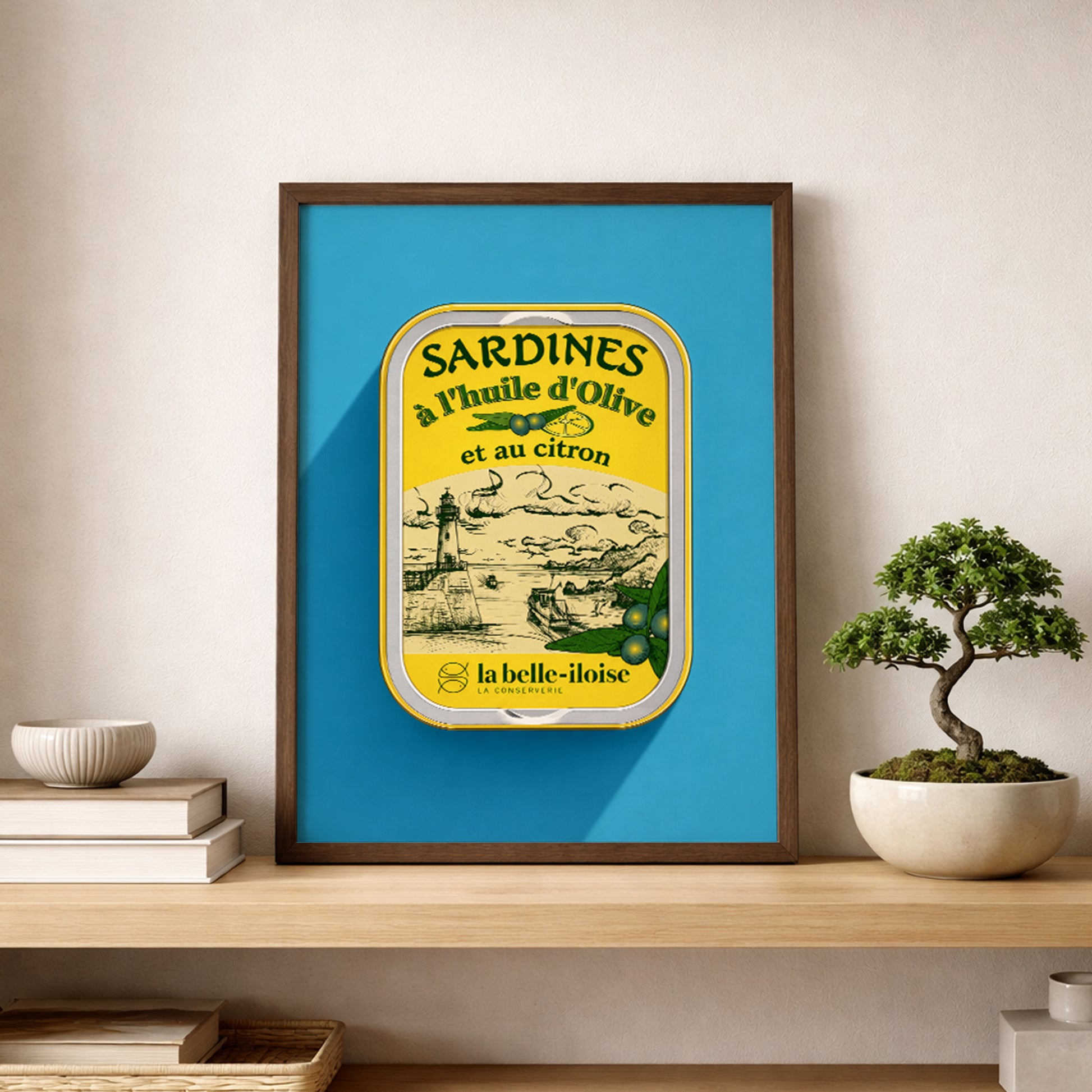 Framed French sardine tin pop art print resting on a wooden shelf beside a small bonsai plant, featuring turquoise blue and yellow vintage packaging artwork in a clean neutral interior.