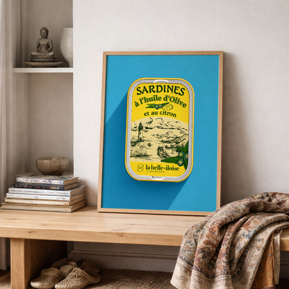 French sardine tin art print leaning on a shelf beside folded patterned textiles, featuring yellow vintage food packaging against a turquoise blue background in a warm, collected interior.