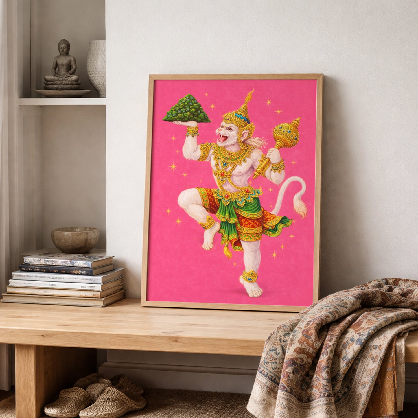 Thai Hanuman Ramakien art print leaning on a shelf with stacked books and textured cultural fabrics. The vivid pink spiritual poster highlights the Hindu monkey god in traditional Thai attire with gold detailing. A mythology-inspired wall decor piece that adds bold colour and Southeast Asian heritage influence to a relaxed interior space.