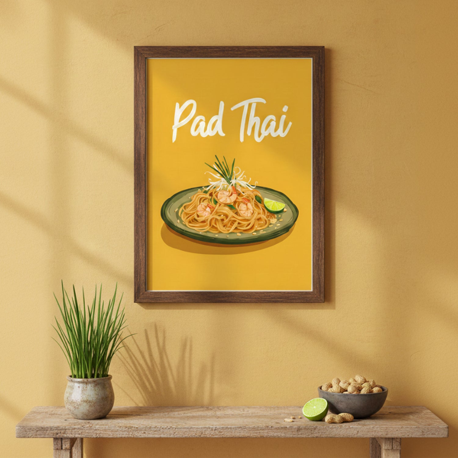 Framed kitchen poster on a warm yellow wall featuring an illustration of a plate of Pad Thai on a green dish with prawns, noodles, bean sprouts, chives and a lime wedge. The words “Pad Thai” appear in white script at the top against a mustard-yellow background. Below the frame is a wooden shelf holding a small potted grass plant, a bowl of peanuts and a halved lime.