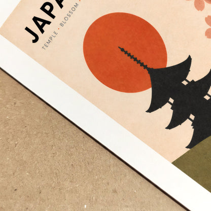 Close-up view of the vintage Japan travel poster showing the printed surface, warm cream paper, bold black temple silhouette and textured red rising sun. Part of the JAPAN title and pink cherry blossom design can be seen at an angle, highlighting the colour, clean edges and tactile finish of this Japanese art print.