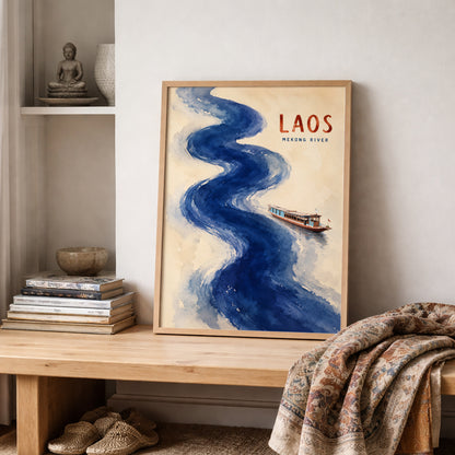 Laos Mekong River poster leaning on a wooden shelf in a cosy neutral room. The cream artwork features a sweeping blue watercolour river, a small traditional boat and red LAOS text, surrounded by soft furnishings, shelving and warm natural decor. Southeast Asian cultural wall art styled for a relaxed living space or bedroom.