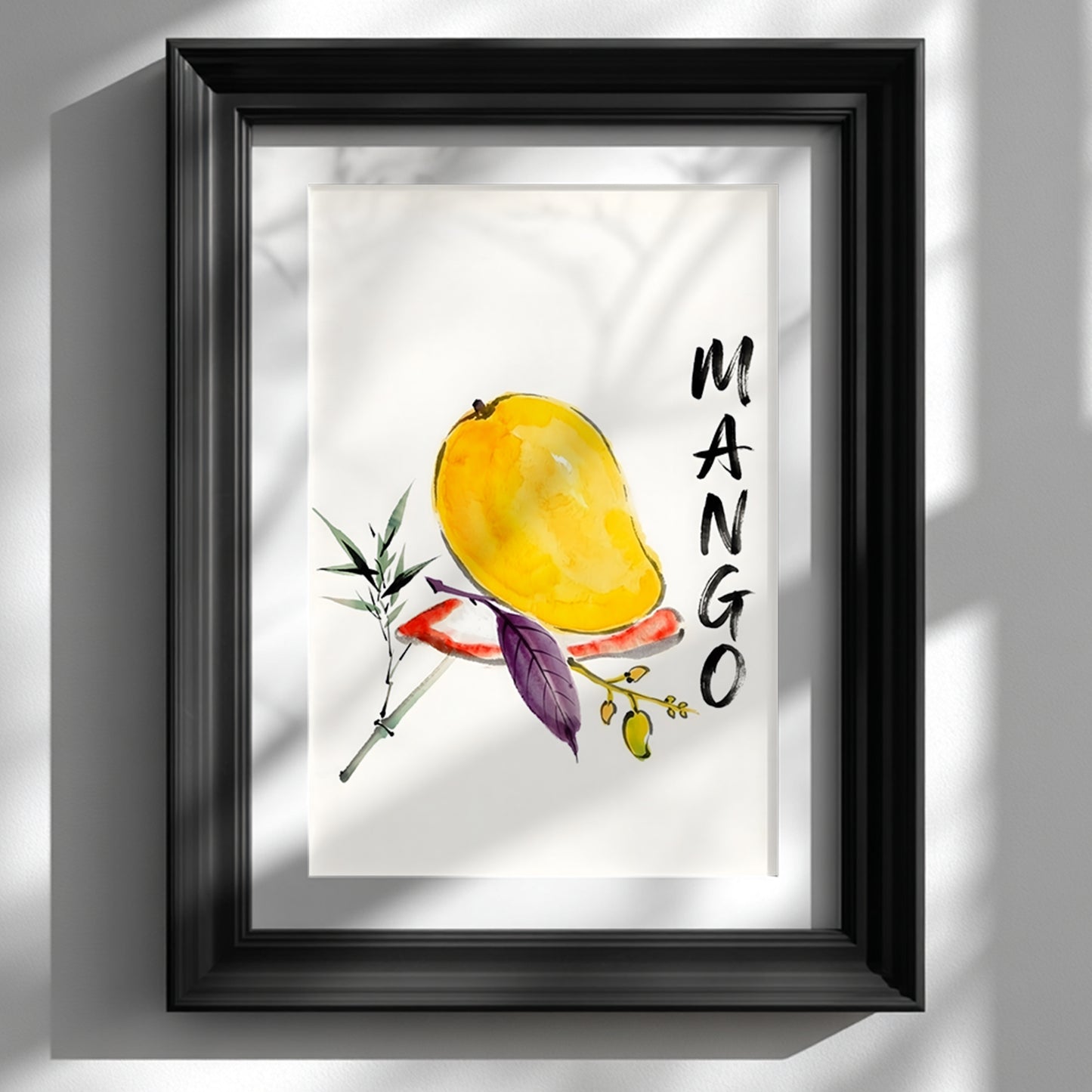 Framed mango wall art print shown on a white wall in a black frame with a white mount. The artwork features a yellow mango painted in loose watercolour with green and purple leaves, a small fruit branch and bold vertical MANGO lettering. Soft window shadows fall across the wall and frame, giving the tropical fruit print a clean, modern look suited to kitchen wall art or bright dining spaces.