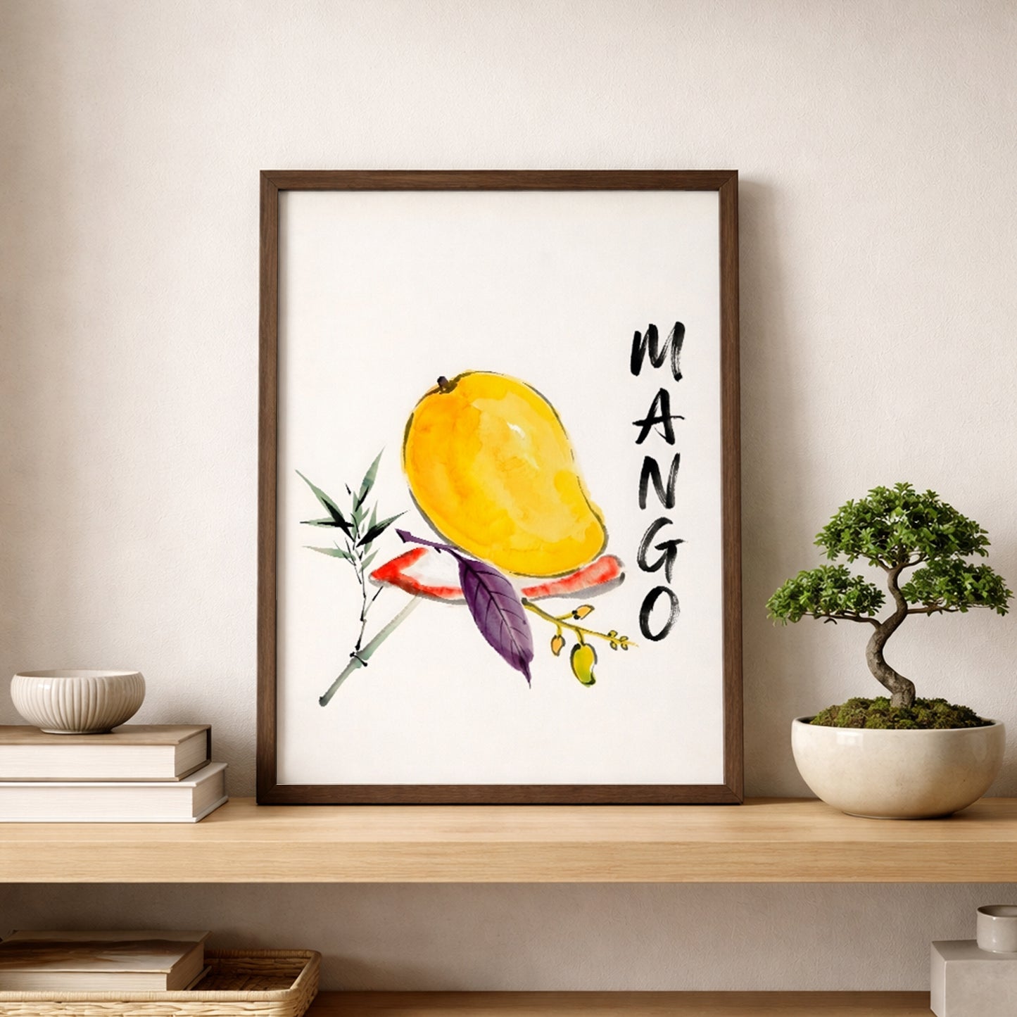 Framed mango print styled on a wooden shelf against a soft neutral wall, placed beside stacked books and a small bonsai-style plant. The artwork shows a yellow mango painted in watercolour with green and purple leaves, a small fruit branch and vertical MANGO text. The shelf styling presents the piece as modern kitchen wall art or tropical fruit wall art, with a simple botanical look that suits calm, bright interiors.