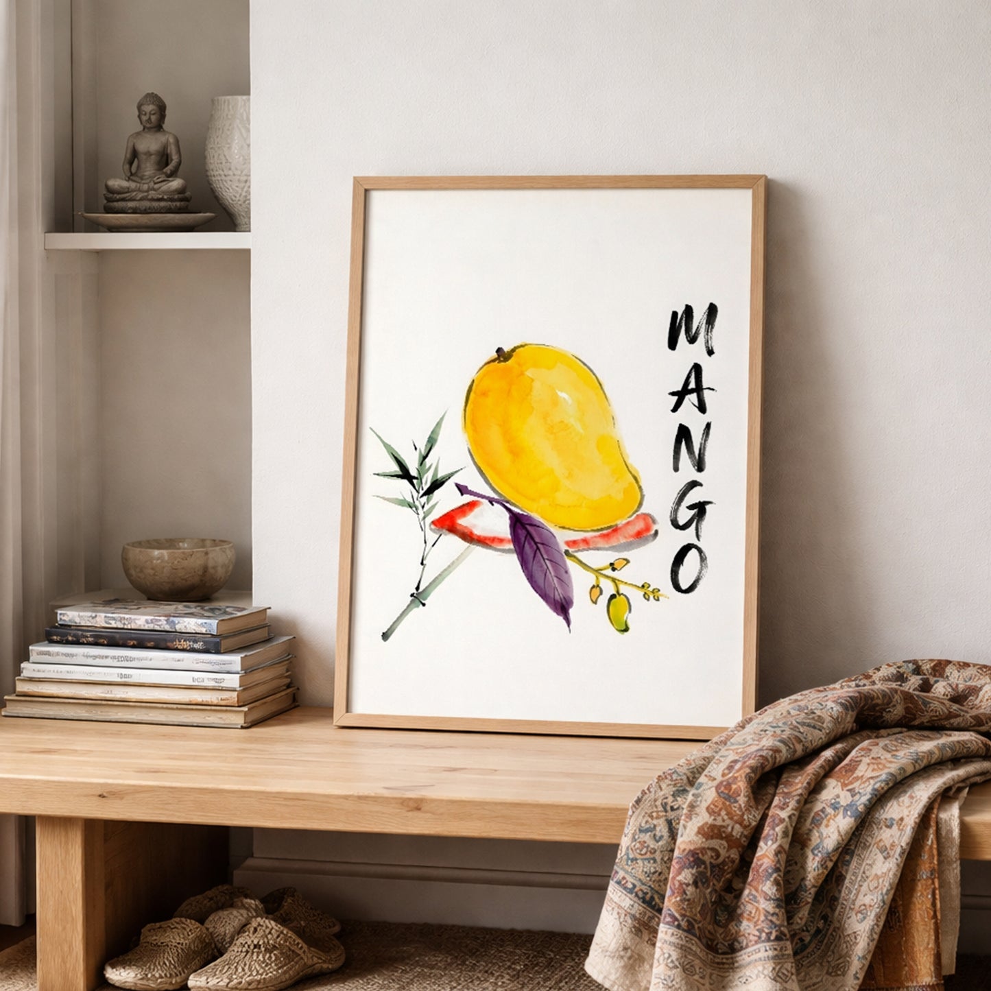 Mango art print in a light wood frame displayed on a shelf in a cosy neutral room with books, pottery, a small plant and folded textured fabrics. The artwork features a yellow mango in loose watercolour, green and purple leaves, a small fruit branch and bold vertical MANGO text. This lifestyle image shows the tropical fruit print as relaxed botanical wall art for kitchens, dining rooms or warm modern interiors.
