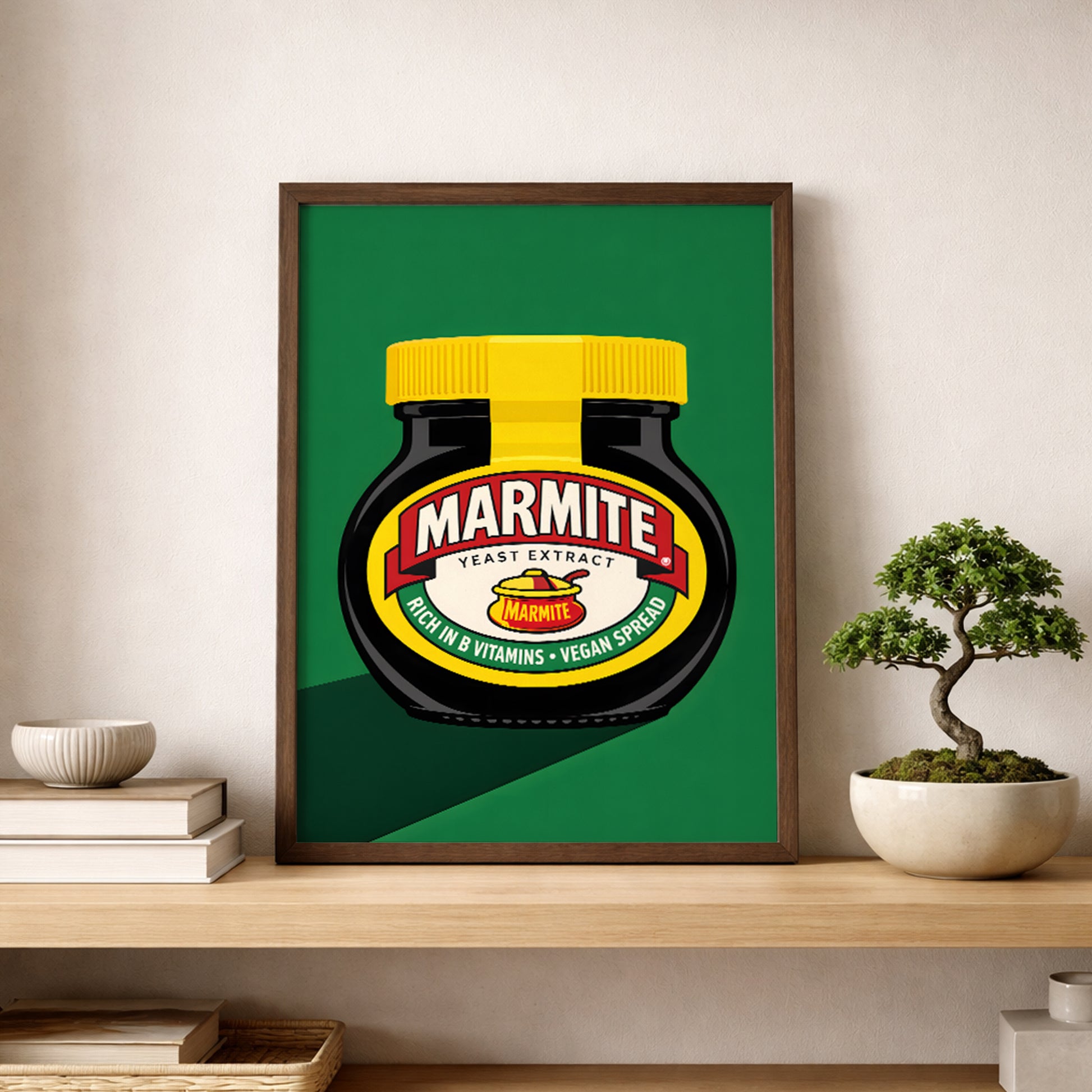 Marmite Jar Print leaning on a shelf against a neutral wall beside a small bonsai-style plant and stacked books. The artwork features a black Marmite jar with a yellow lid, red and cream label, and green background in a bold graphic layout. The simple shelf styling shows how the print works as contemporary kitchen wall art, British pop art decor or modern food-themed shelf display.