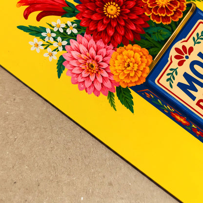 Close-up angled view of the Mexican mole poblano print resting on a textured surface, showing the yellow background, vivid flower petals and blue vintage tin label in sharp detail. The crop highlights the printed colour, paper surface and hand-painted packaging style of this bright Mexico kitchen wall art.