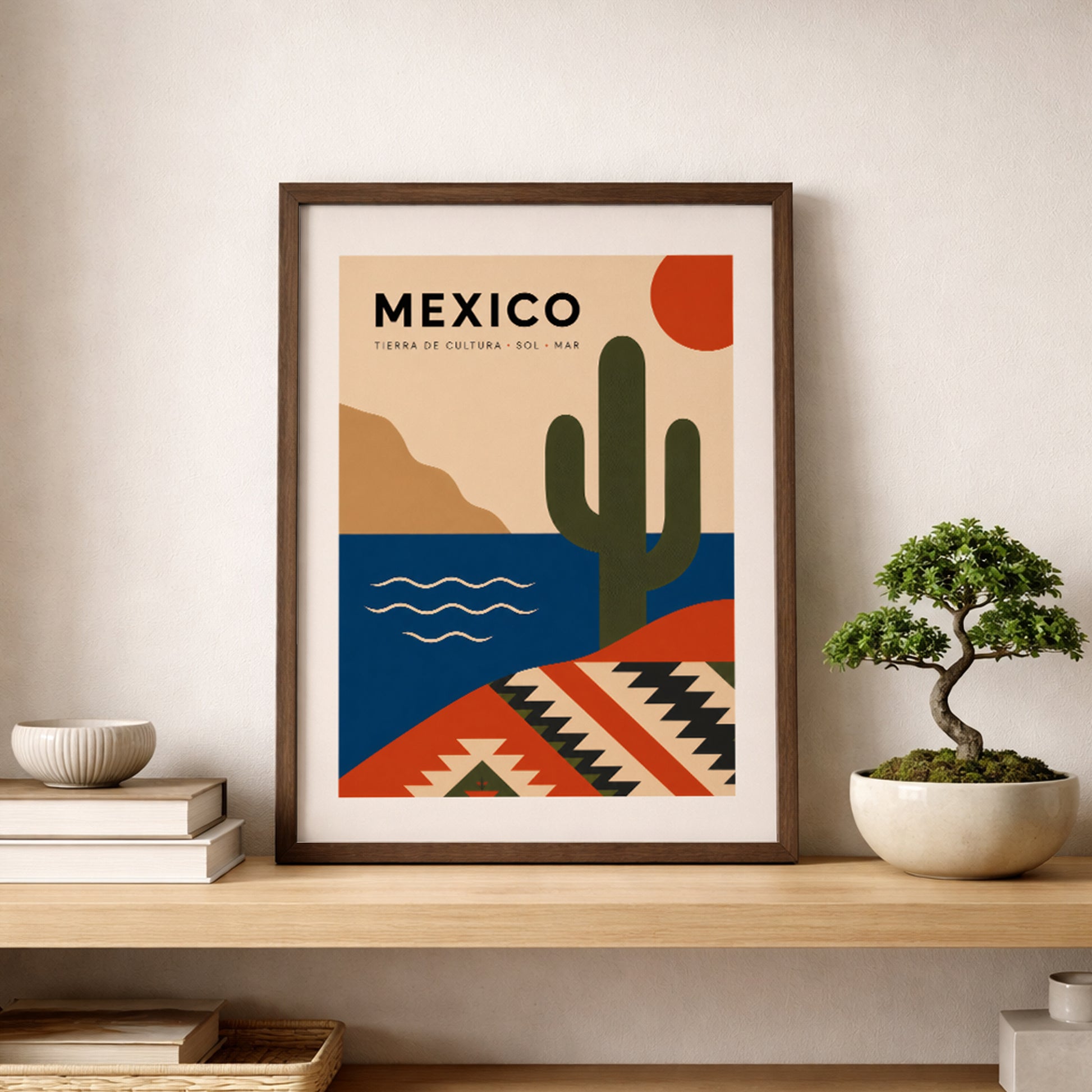 Natural wood framed Mexico travel poster resting on a shelf beside stacked books and a small bonsai tree. The artwork features a green cactus, red sun, blue sea and geometric Mexican textile pattern, creating a warm cultural wall art display for a living room, bedroom, study or reading corner.