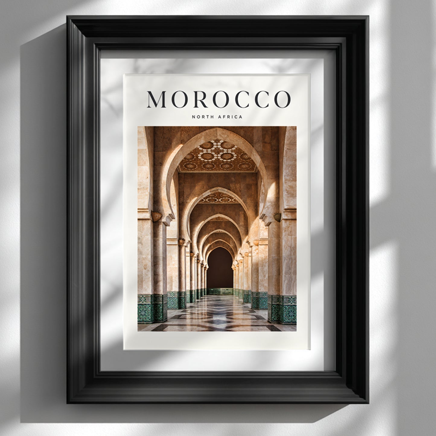 Morocco travel print showing the arched architecture of Hassan II Mosque in Casablanca, displayed in a black frame on a white wall with natural sunlight and shadows.