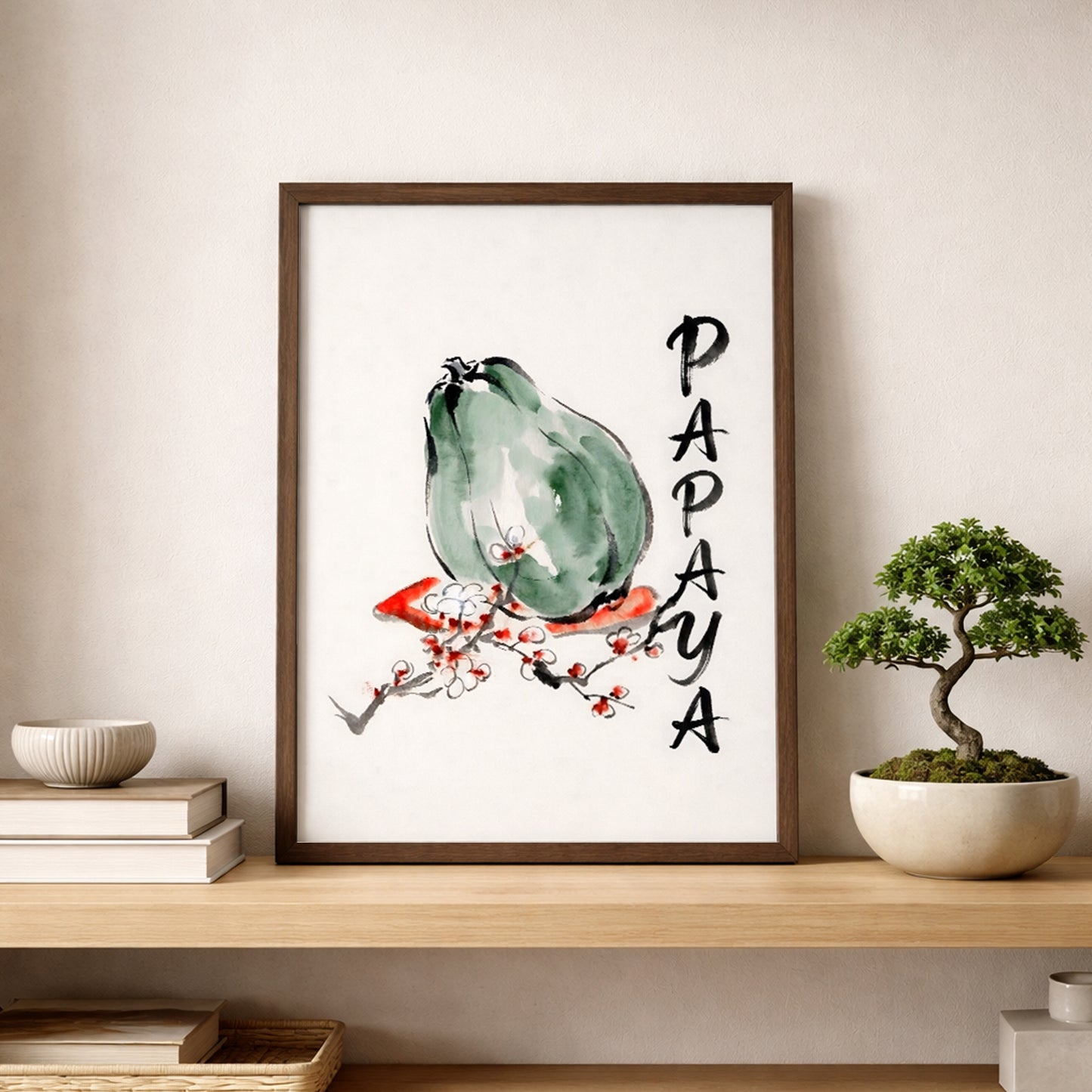 Framed papaya print displayed on a shelf against a light neutral wall beside stacked books and a small bonsai style plant. The artwork shows a green papaya in loose watercolour and ink, with a red blossom branch and vertical PAPAYA text. Lifestyle image for tropical fruit wall art, papaya kitchen print, botanical decor, and modern minimalist shelf styling.