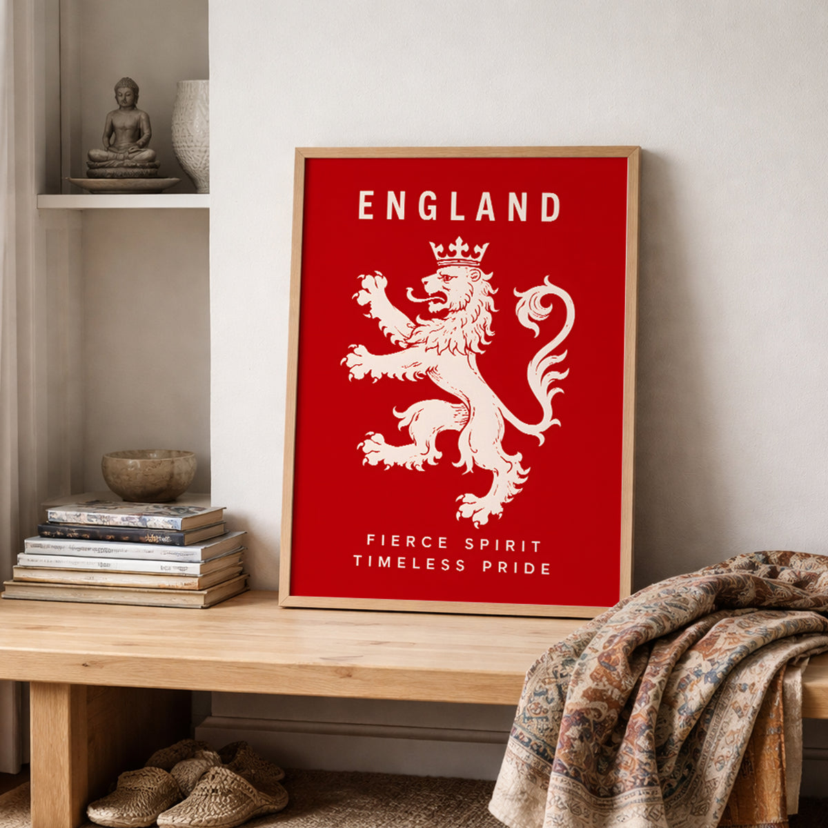 A framed red England poster featuring a white lion rampant and the words 'ENGLAND' and 'FIERCE SPIRIT TIMELESS PRIDE' leans against a wall on a wooden bench in a styled room with books, a blanket, and woven slippers.