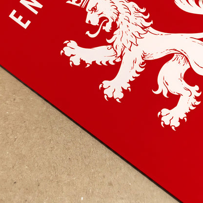 A close-up of a red rectangular product, likely a mat or board, featuring a white lion rampant graphic and partial white lettering, placed on a tan textured surface.