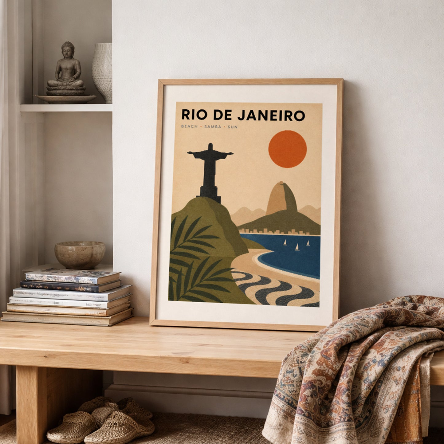 Framed Rio de Janeiro travel poster resting on a shelf beside stacked books, pottery and a textured woven throw. The vintage-style Brazil print features Christ the Redeemer, Sugarloaf Mountain, Copacabana Beach and tropical plants, adding warm orange, green and blue colour to a cosy neutral room.