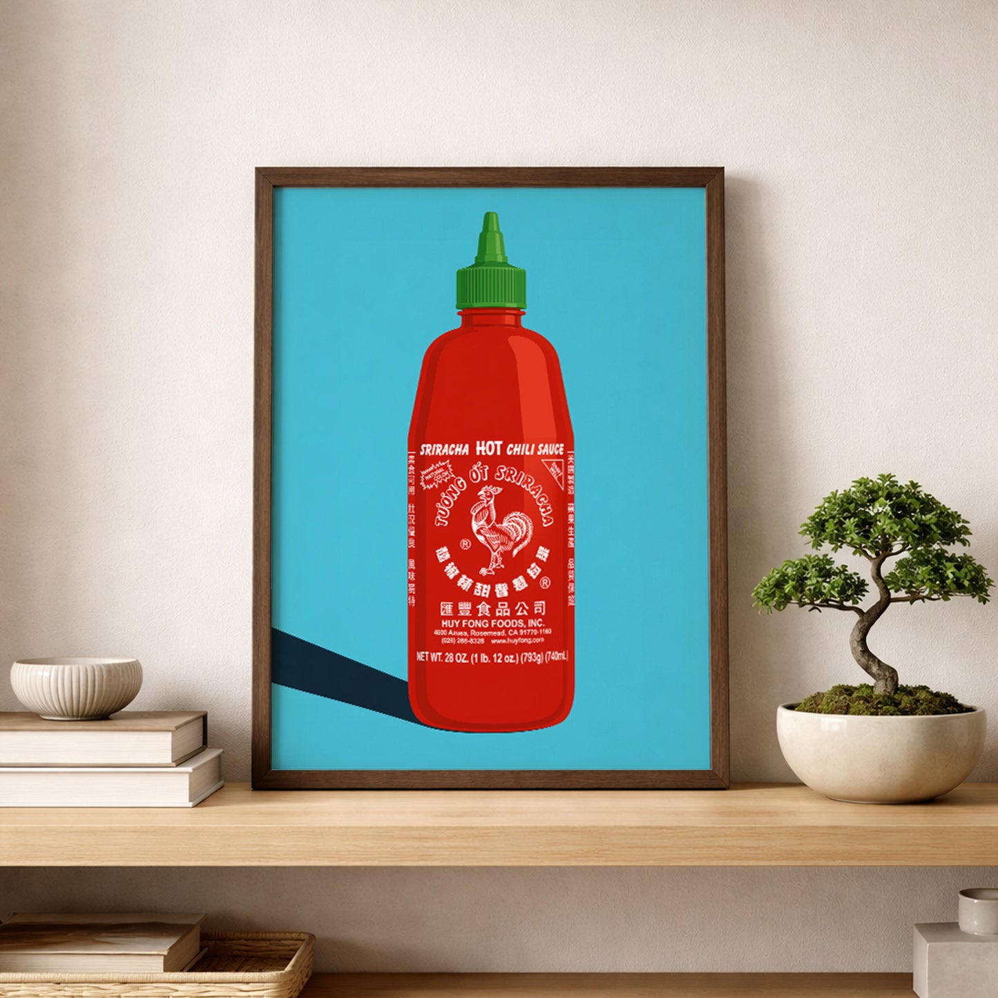 Framed sriracha poster displayed on a simple shelf beside a small bonsai plant and neutral decor. The red hot sauce bottle, green cap, rooster label and bright blue background create a clean pop art kitchen print, ideal for modern kitchen wall decor, dining room wall art, cafe styling or foodie gift ideas.