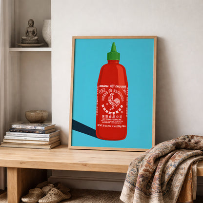 Sriracha wall art displayed on a shelf with books and soft home styling, featuring a red hot sauce bottle, green cap, white rooster logo and blue background. A bold food poster for kitchen wall decor, dining rooms, cafes, restaurant walls or anyone looking for a colourful spicy food print.