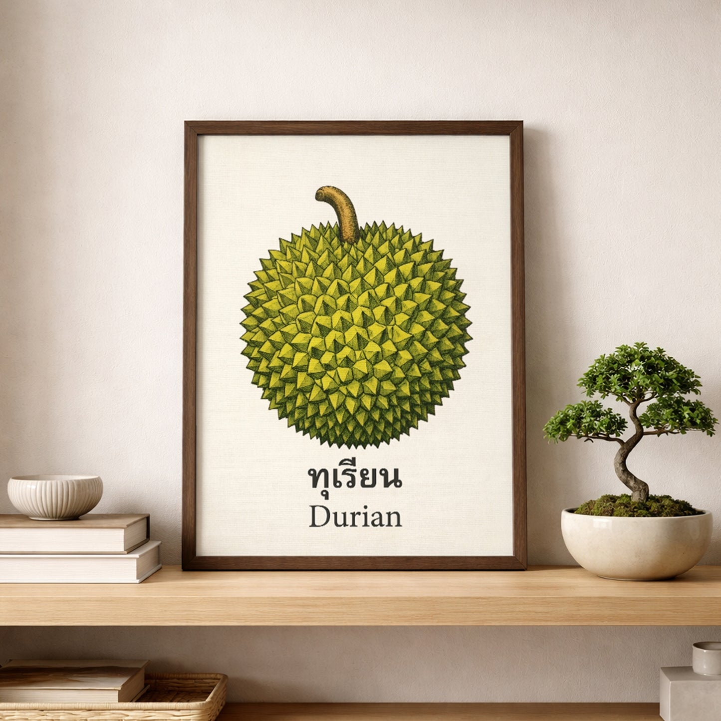 Framed Thai durian poster styled on a shelf beside a small bonsai plant in a calm neutral interior. The print features a green durian fruit with a brown stem, Thai lettering and an English label on a cream background. The scene shows how the artwork works as minimalist kitchen wall art, dining room decor, cafe styling or a simple tropical fruit print in modern spaces.