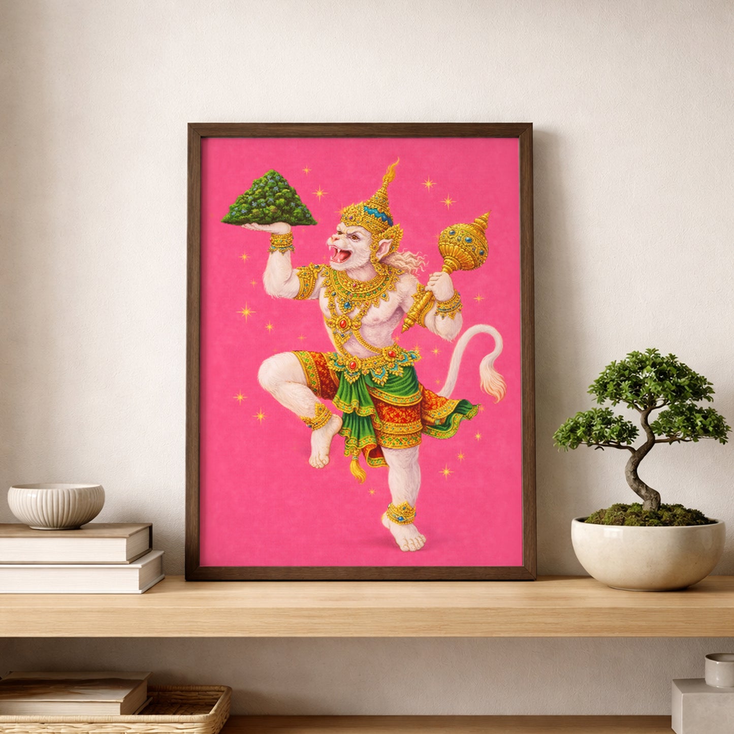 Thai Hanuman Ramakien art print styled on a wooden shelf beside a bonsai plant and minimal decor. The vibrant pink mythology poster features the Hindu monkey god in ornate Thai costume, creating a bold focal point within a calm, natural interior setting. A spiritual wall art piece combining Southeast Asian folklore with contemporary home styling.