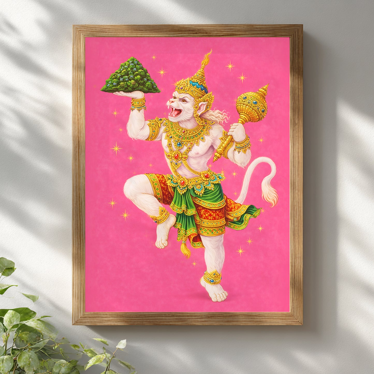 Thai Hanuman Ramakien art print displayed in a natural wooden frame against a white wall with soft light and plant shadows. The vibrant pink background enhances the detailed depiction of the Hindu monkey god in traditional Thai costume. A spiritual mythology wall decor piece combining Ramakien folklore with modern minimalist interior design.