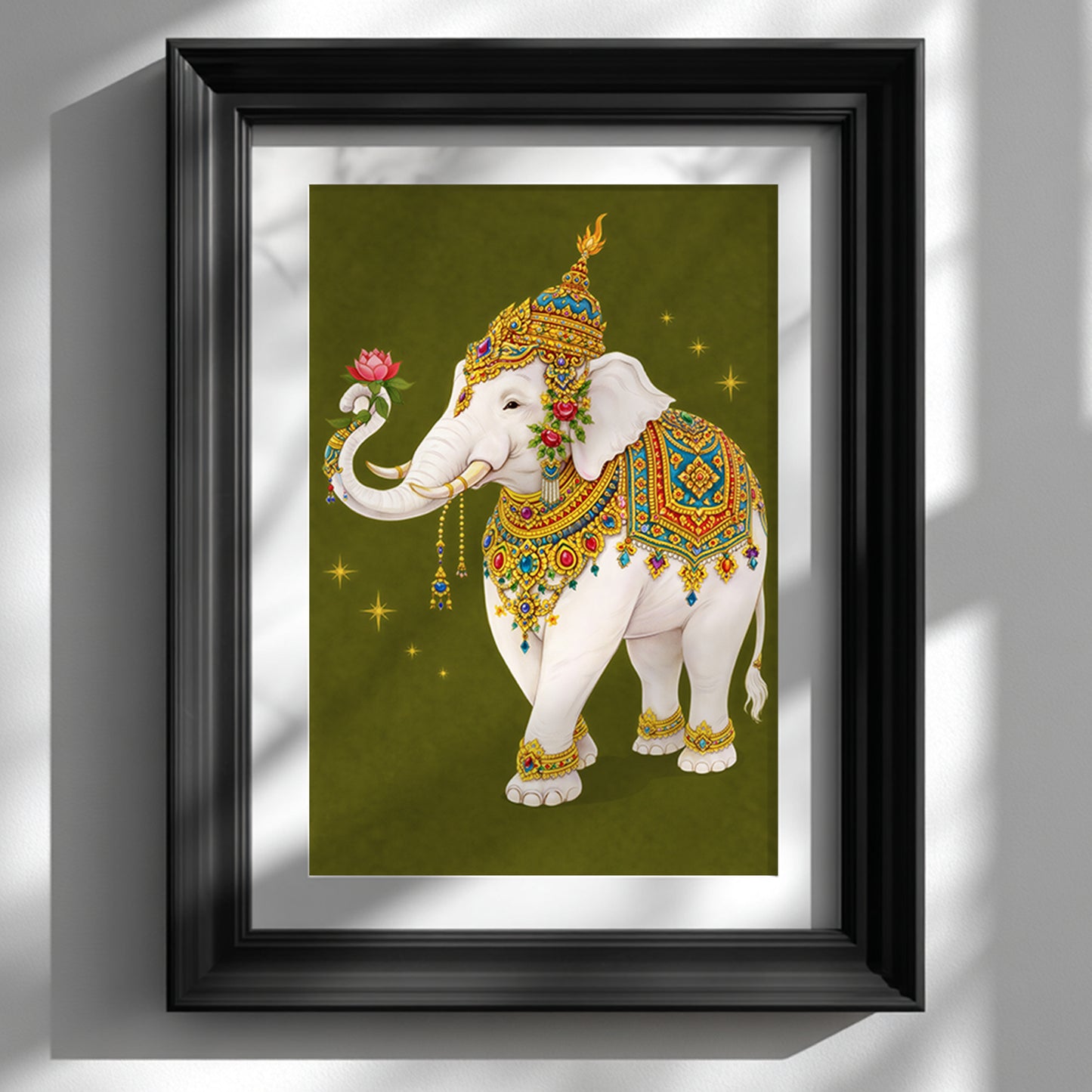 Thai royal white elephant wall art displayed in a black frame against a white wall. The sacred white elephant is illustrated in ornate Thai ceremonial regalia with gold, red and turquoise detailing, holding a pink lotus flower in its trunk. The olive green background with subtle star accents enhances the traditional Southeast Asian aesthetic in a refined, gallery-style interior setting.