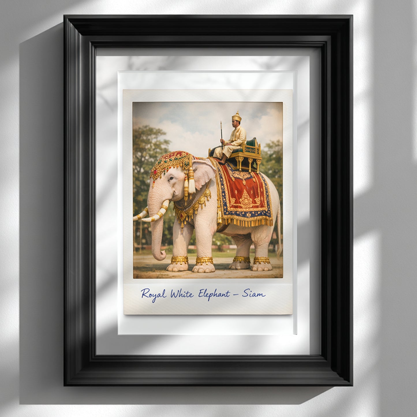 Framed Thailand royal white elephant poster displayed on a white wall in a black frame. The artwork shows a decorated white elephant wearing red and gold royal cloth with gold ornaments, carrying a Thai royal figure in traditional dress. Styled as a vintage Polaroid with handwritten caption, this Thai cultural wall art suits classic and heritage interiors.