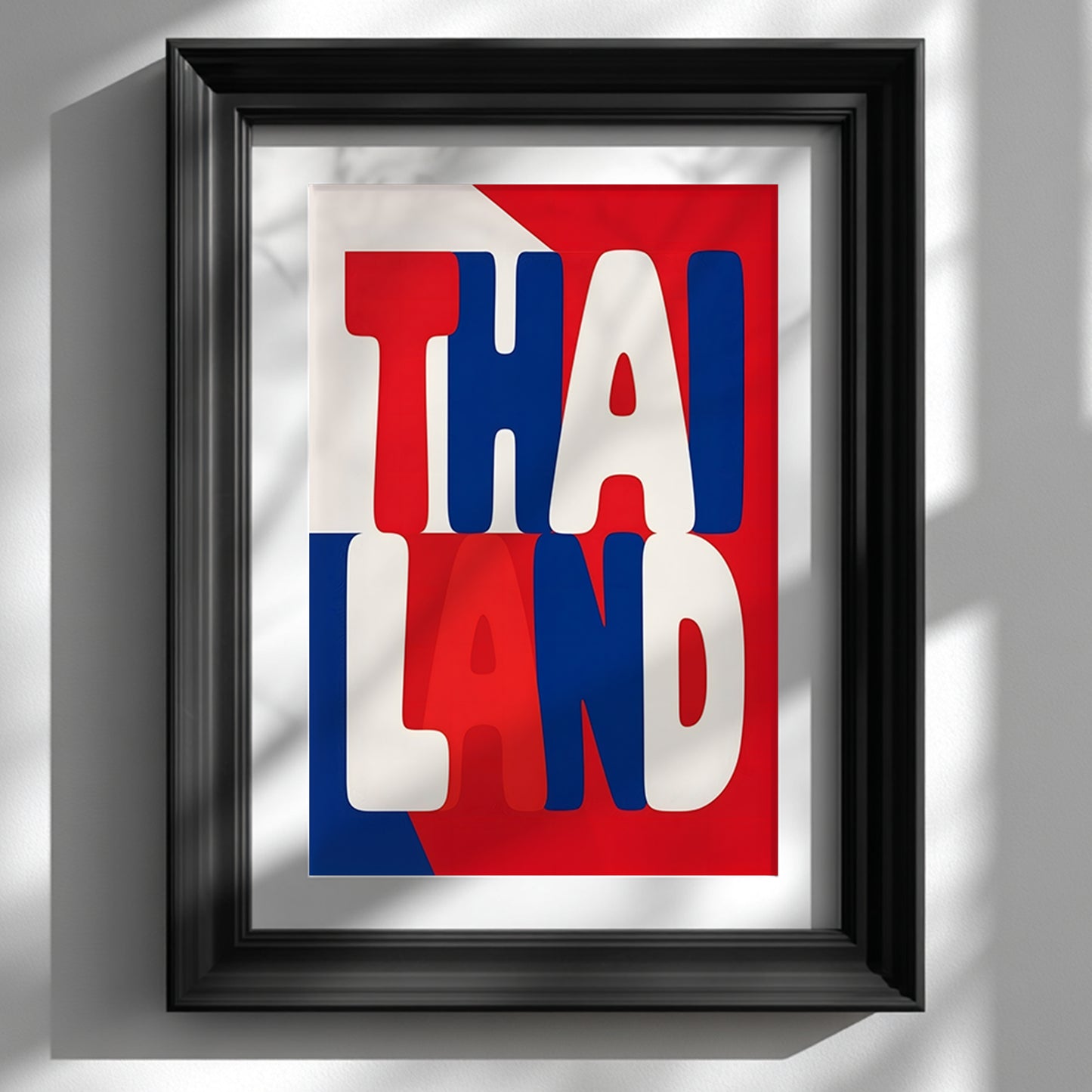 Black-framed Thailand typography poster on a white wall, showing stacked red, white and blue lettering based on Thai flag colours, lit by sunlight and soft leaf shadows.