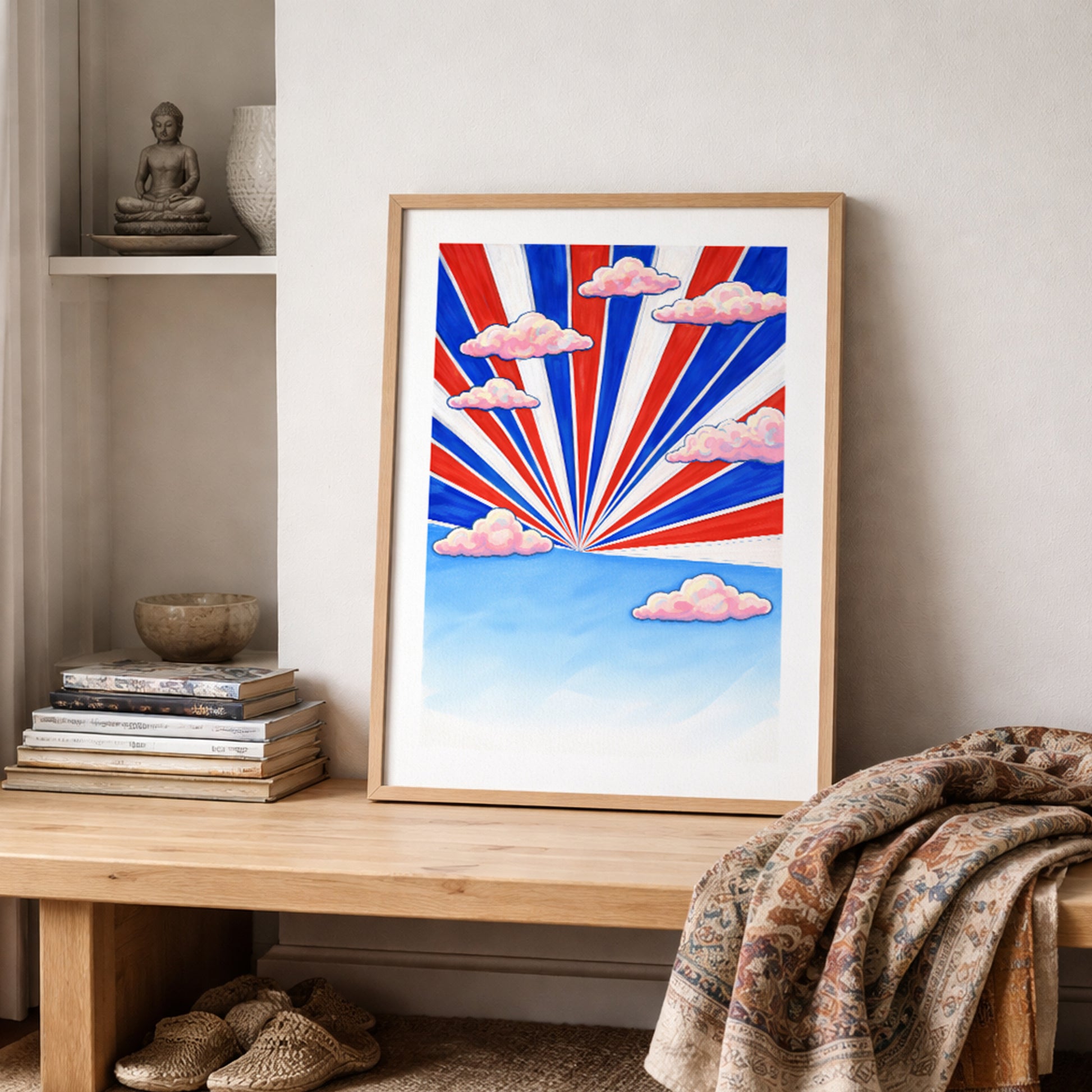 Thailand wall art poster with red, white and blue sunburst rays leaning on a shelf with books and textured decor. Soft pink clouds float over a blue sky horizon. Graphic Thai flag inspired travel print styled for modern home decor and Southeast Asia themed interiors.