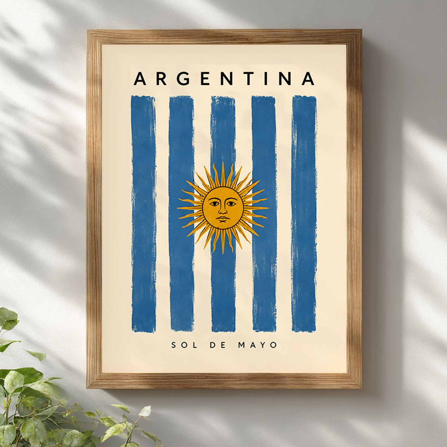 Argentina flag poster displayed in a warm wooden frame against a bright white wall with soft leafy shadows. The artwork features blue and cream stripes, the golden Sun of May, Argentina lettering and small Sol de Mayo text. The natural frame and light interior styling complement this vintage-inspired Argentine travel print.